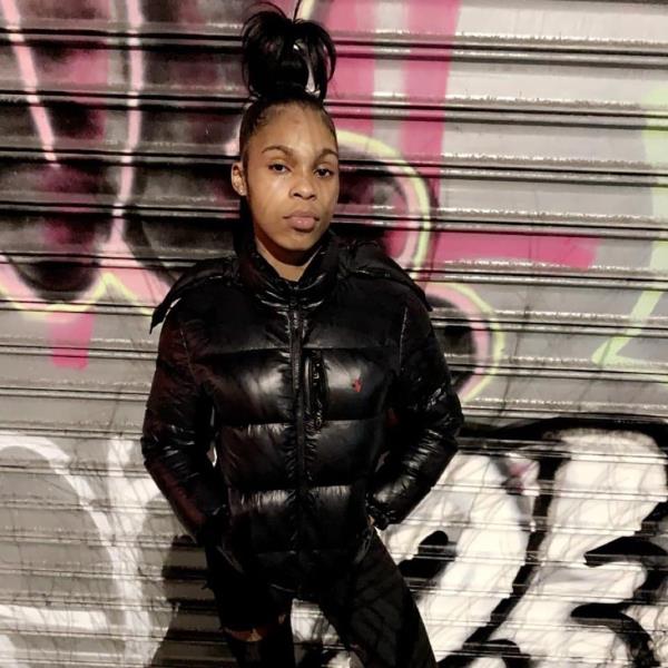 Keyaria Rattray-Brother stands infront of a graffiti covered rollup gate wearing all black