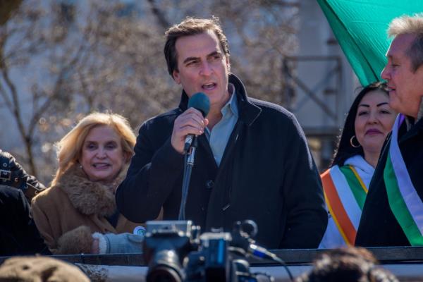 Michael Gianaris speaking at an outdoor event with former Rep. Carolyn Malo<em></em>ney in the background with several other people standing alo<em></em>ngside them. 