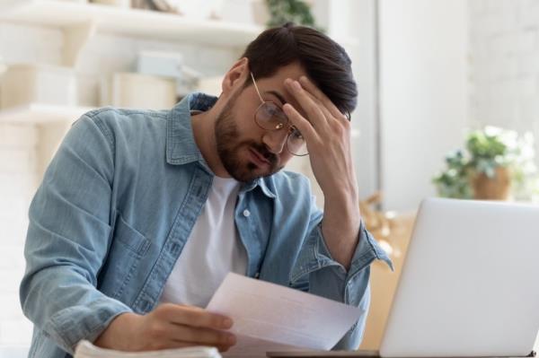 Upset frustrated young man reading bad news in postal mail letter paper docu<em></em>ment sit at home table, depressed stressed guy worried a<em></em>bout high bill tax invoice, overdue debt notification mo<em></em>ney problem
