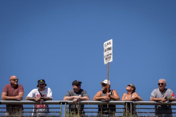 A line-up of men lean against a railing. One of the men is holding a protest sign.